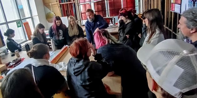 Mehrere Personen sitzen und stehen um einen großen Holztisch in einer Werkstatt mit großen Fenstern und bunten Glasmalereien an der Wand.